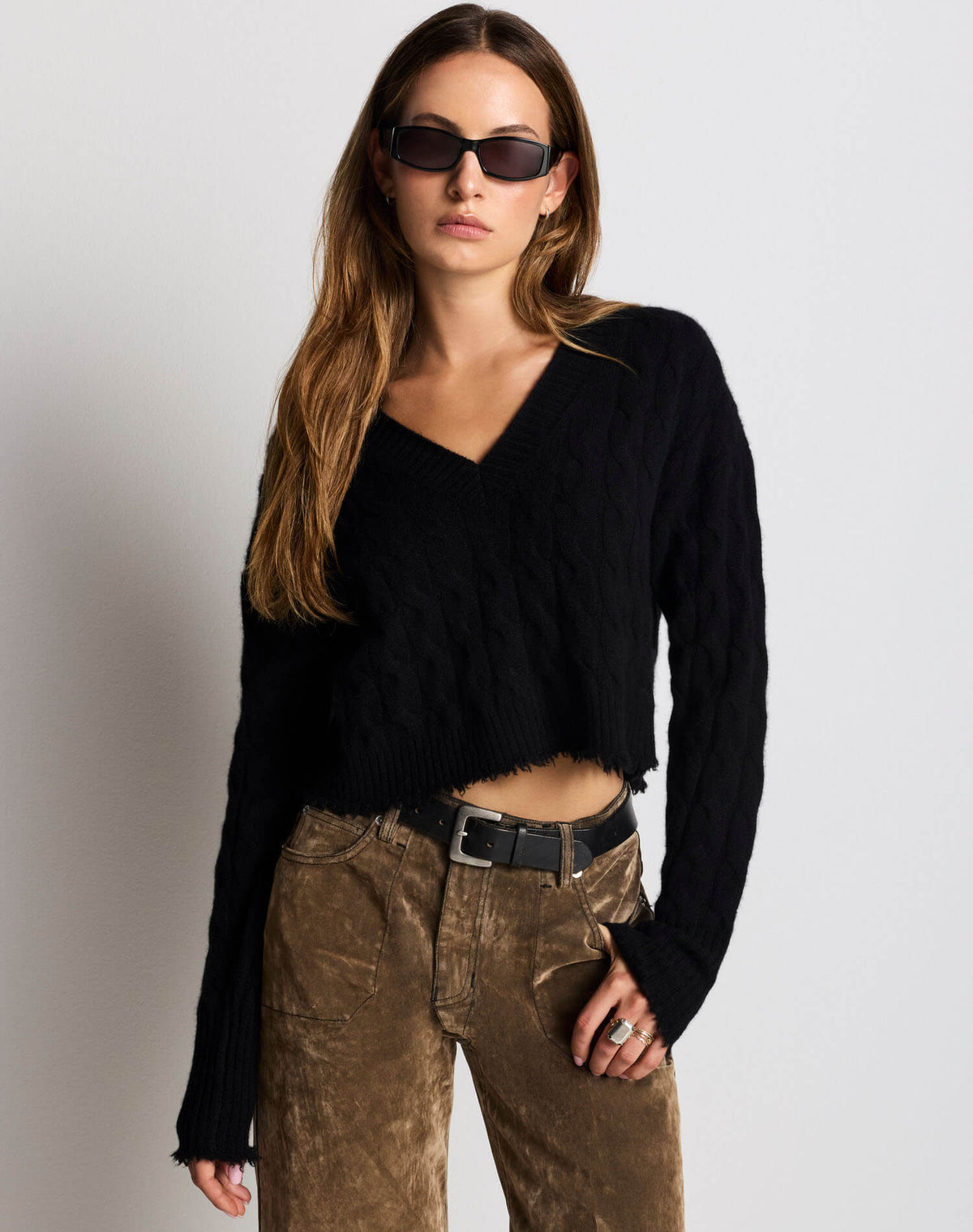 A woman modeling a black long-sleeve cropped V-neck knit sweater with frayed edges, paired with brown corduroy pants and black sunglasses, against a light gray studio background.