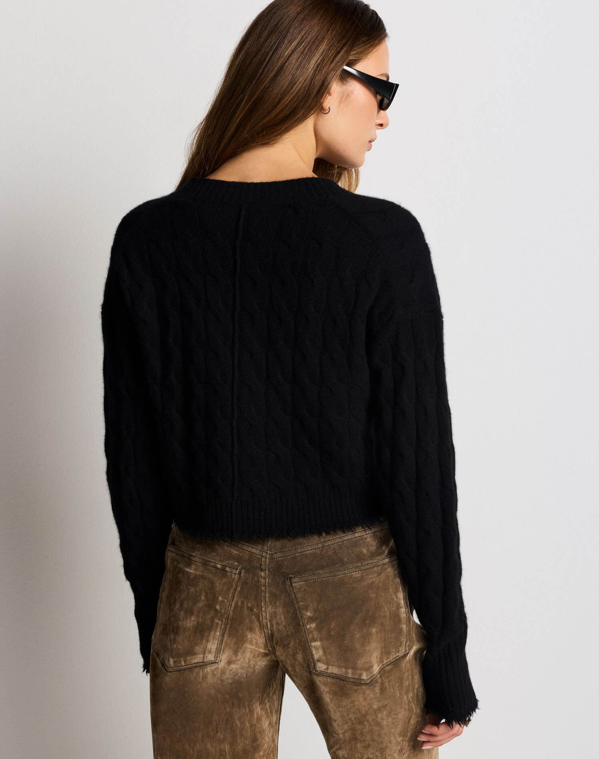 A woman shown from the back wearing a black cable-knit cropped sweater and brown jeans against a plain light gray studio background.