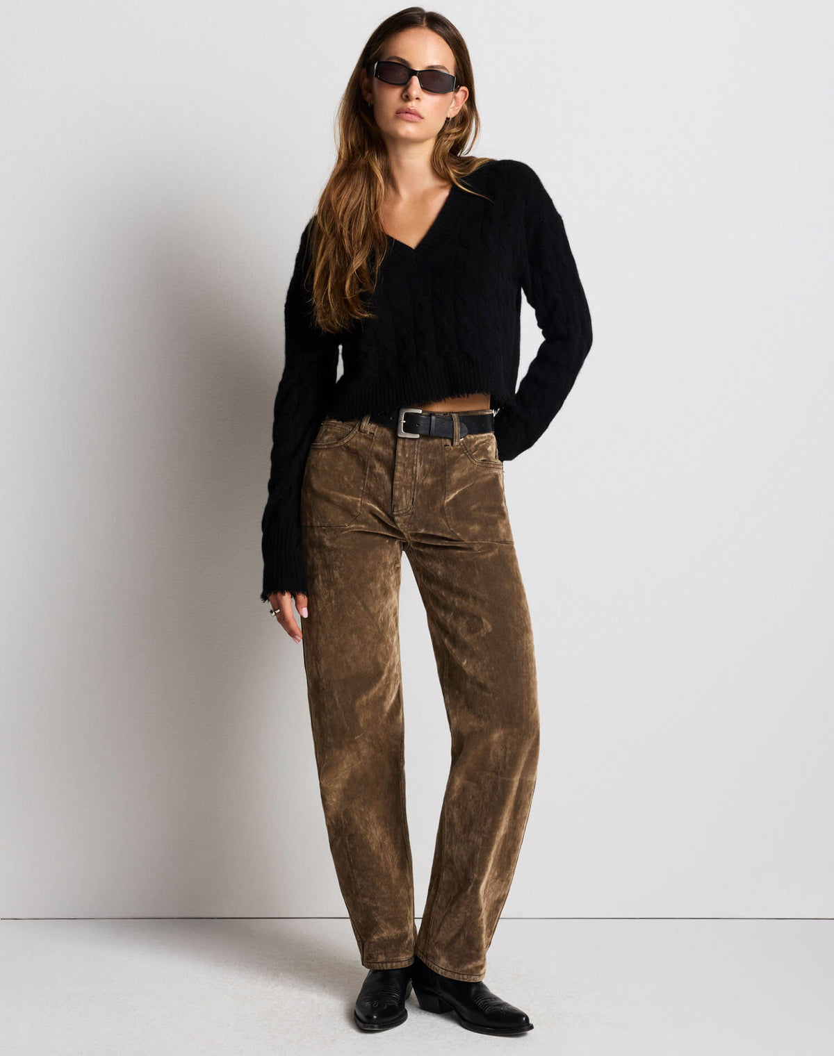 A woman modeling a black long-sleeve cropped V-neck top and brown corduroy pants with a black belt, styled with black sunglasses and black boots against a plain light gray studio background.